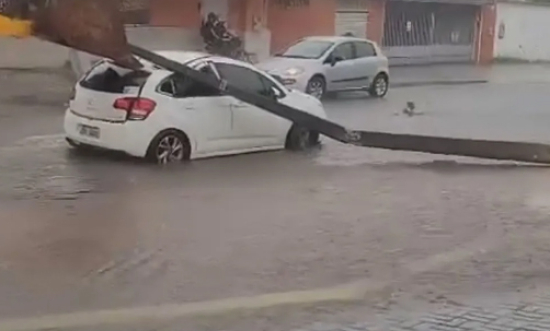 Poste cai em cima de carro que passa por rua durante chuva